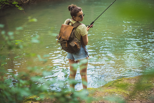 View of a young woman fishing in a calm stream. She has her backpack on in order to be light on feet and change position whenever she feels like the fish is not biting. Room for copy space.
