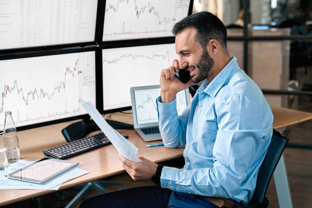 istock Stock Traiding. Trader sitting in front of monitors with data at office talking on phone doing online trading training reading document smiling cheerful 1271844023