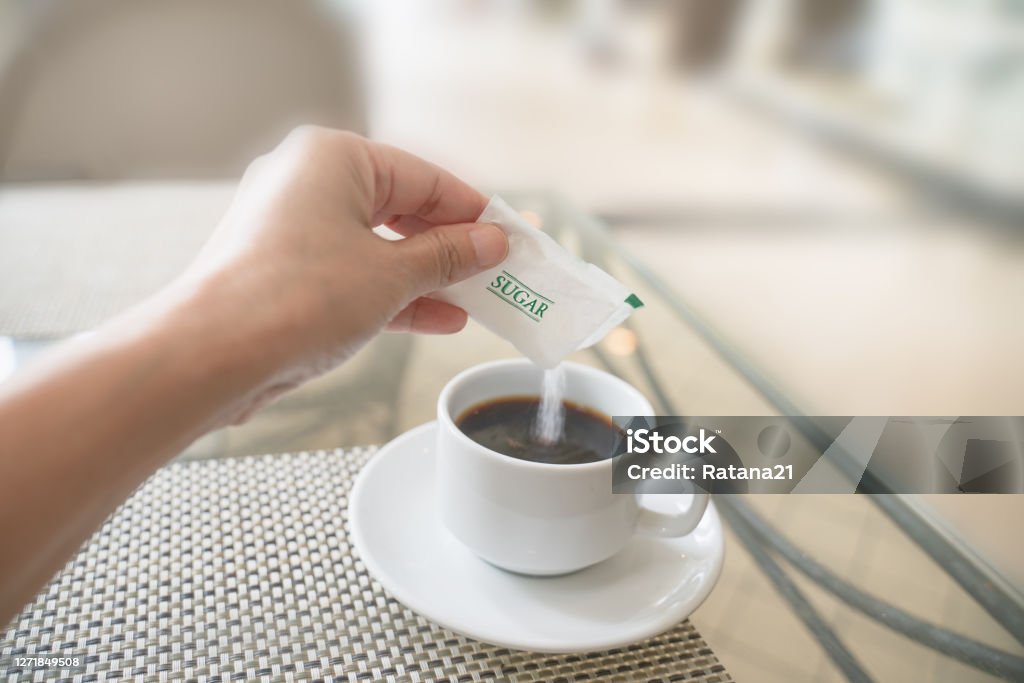 human hand with white sugar packet and pouring in hot black coffee , in soft focus Sugar - Food Stock Photo