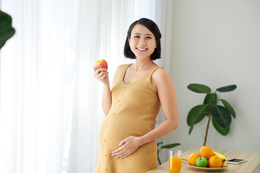 Pregnant woman standing near table with plate of fresh fruits indoors