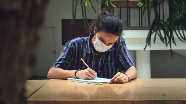 Front view of a masked young university student studying alone in the library stock photo