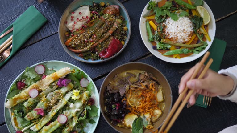 Top view of people eating their healthy vegan meals with chopsticks. Bowls full of colorful vegetables served on black chunky wooden table