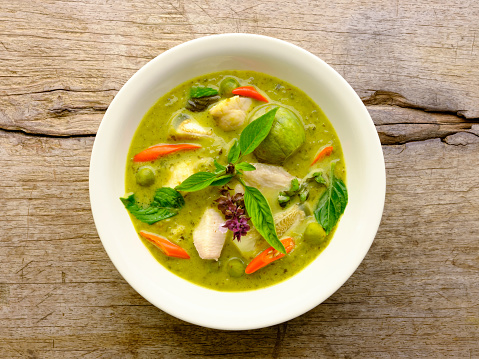 Famous internationally renowned Thai green coconut curry 'Gaeng Keow Wan Gai', with chicken, in a white bowl, sitting on an old worn wood table background.