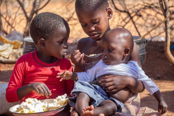 three african children in a village near Kalahari desert three african children in a village near Kalahari desert, the sister feeding her brother in the outdoors kitchen developing countries photos stock pictures, royalty-free photos & images