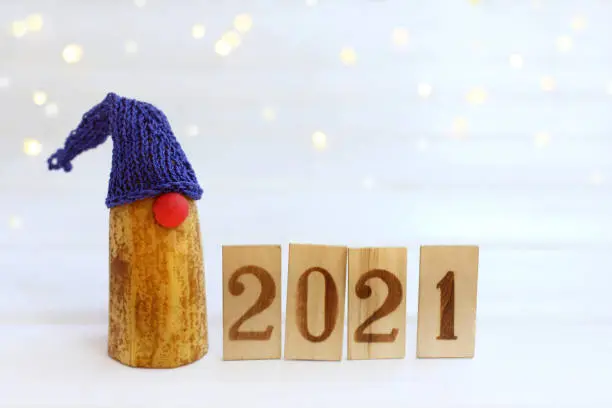 funny gnome in a blue hat with wooden numbers 2021 on a background of festive lights