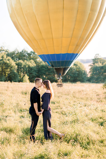 Couple in love stands face to face, holding hands, in summer field on the background of yellow air balloon. Man and woman in black clothes, celebrating their anniversary with flying on balloon.