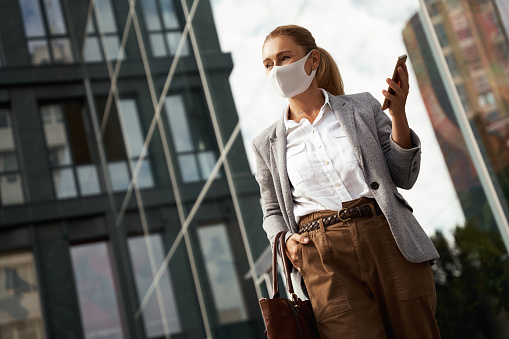 Social distancing. Confident business woman wearing protective face mask holding mobile phone while waiting for partner, standing near office building outdoors. Covid 19, coronavirus and business