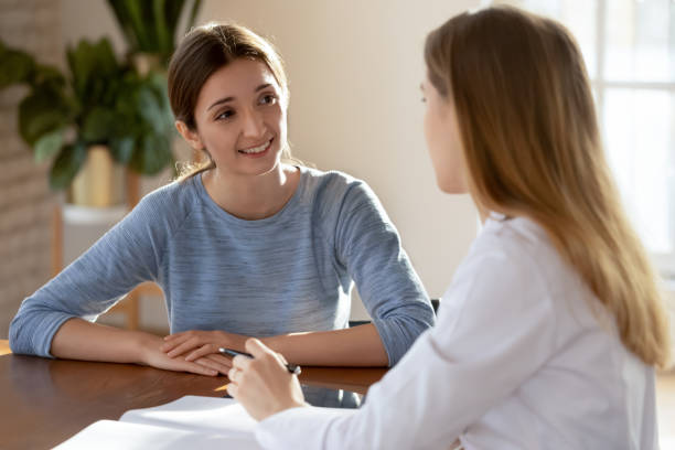 Smiling young woman patient listening to doctor at meeting Smiling young woman listening to doctor therapist at meeting, sitting at work table in hospital, physician gp consulting patient about checkup results, giving recommendations at medical appointment general view stock pictures, royalty-free photos & images