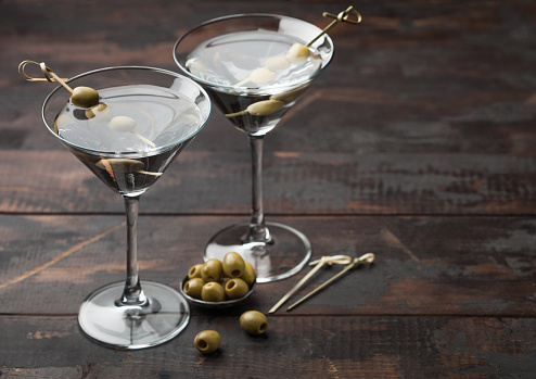 Vodka martini gin cocktail in original glasses with olives in metal bowl and bamboo sticks on dark wooden background.
