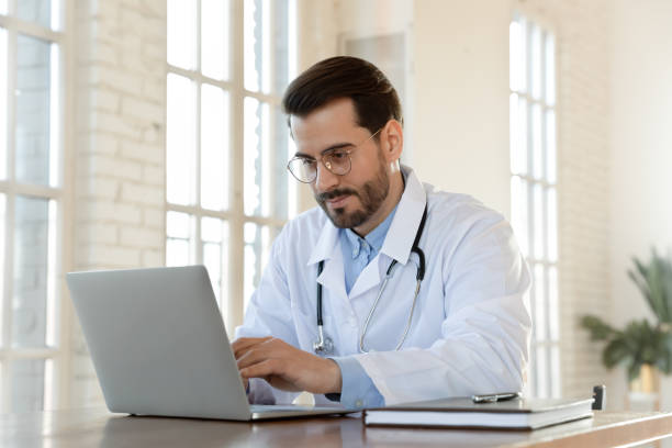 giovane medico concentrato che indossa occhiali che lavorano su laptop in ufficio - trainee computer medical student student foto e immagini stock