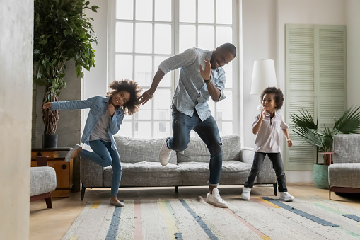 Happy african american father showing funny dancing moves to energetic small children siblings at home. Laughing crazy little biracial boy and girl having fun with caring daddy in living room.