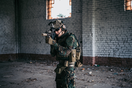 Military soldier in the form holds in the hands an assault rifle, check possible enemies around the corner.
