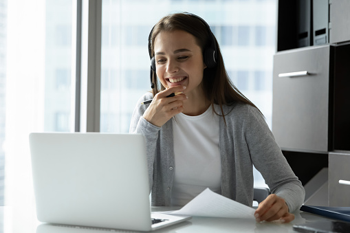 Smiling young woman wearing headset looking at laptop screen, studying or working online, listening to lecture, watching webinar, writing notes, support service employee consulting client