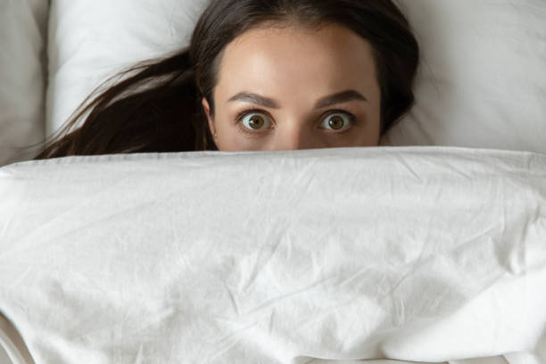 Close up top view funny young woman peeking from duvet Close up top view funny young woman peeking from duvet, scared shocked female looking at camera, lying in bed with wide open eyes, afraid of nightmares, hiding under white warm blanket horror waking up bed women stock pictures, royalty-free photos & images