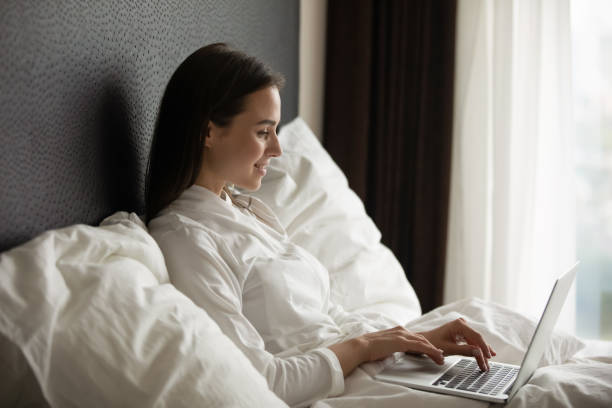 istock Smiling young woman wearing bathrobe using laptop, sitting in bed 1271917101