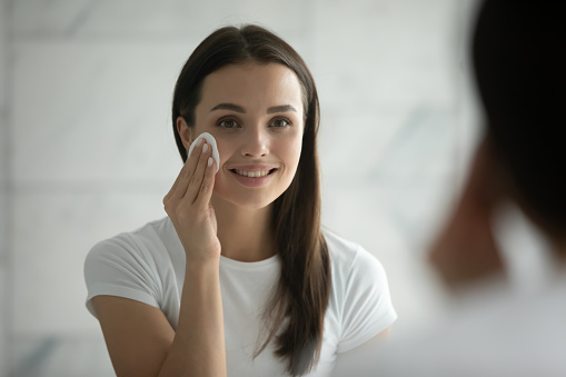 Close up head shot smiling young woman wearing white t-shirt removing face makeup, using cotton pad, standing in bathroom, looking in mirror, cleaning face, enjoying skincare procedure