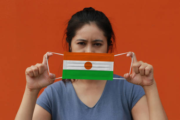 una mujer con bandera de níger en la máscara higiénica en la mano y levantó la cara frontal sobre fondo de color naranja. - arms lifted fotografías e imágenes de stock