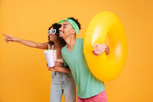 Portrait of a happyyoung african couple in summer clothes holding inflatable ring and pointing finger away isolated over yellow background