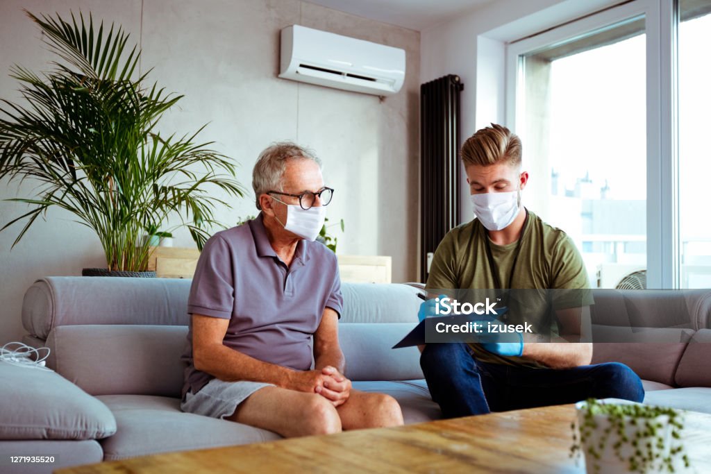 Home caregiver visiting senior man District nurse wearing protective face mask and gloves talking with senior man during home visit. They are sitting in living room. Protective Face Mask Stock Photo