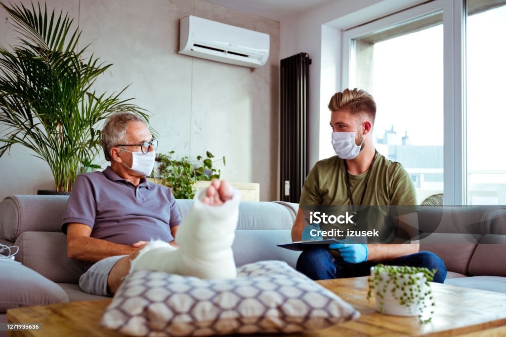 Home caregiver visiting senior man District nurse wearing protective face mask and gloves talking with senior man during home visit. They are sitting in living room. Senior man has broken leg. Broken Leg Stock Photo