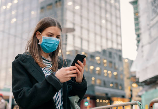 istock Young businesswoman with protective face mask using smartphone while commuting in downtown city street and city buildings with multi-coloured commercial signs at night 1271939649