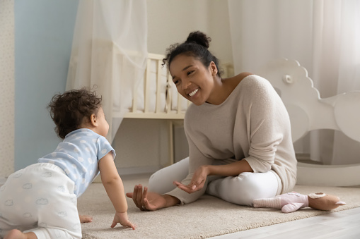 Adorable happy mixed race baby girl or boy creeping, walking on knees and hands on carpet floor to smiling young african american mother, having fun together in children bedroom, childcare concept.