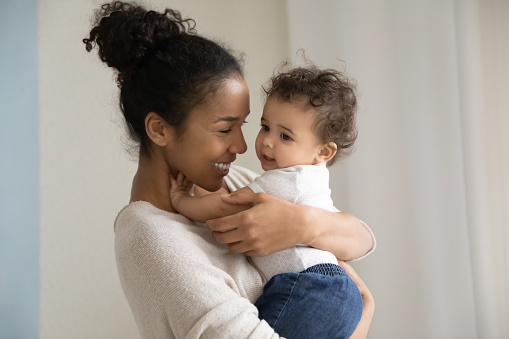 Head shot affectionate millennial biracial mommy or babysitter carrying on hands funny cute curly mixed race kid girl or boy, enjoying daycare time together indoors, responsible parenting concept.