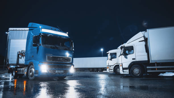 blue semi-truck avec remorque cargo s’éloigne de l’espace de stationnement de nuit où d’autres camions sont debout. le camion long-courrier quitte le parking, transportant des marchandises et des marchandises à travers le continent. rainy night - truck parking horizontal shipping photos et images de collection