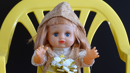 kolenyakh. Na chernom izolyate.\n\nA children's doll with blue eyes, light brown hair in a capishon sits on a yellow chair with a gift on its knees. On black isolate.