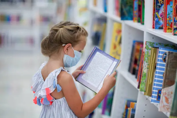 A little girl stands in a bookstore in a medical mask and reads. Child in the library.