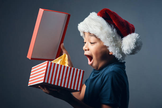 There is a Santa! Studio shot of a cute little boy opening a Christmas present against a grey background unwrapping stock pictures, royalty-free photos & images
