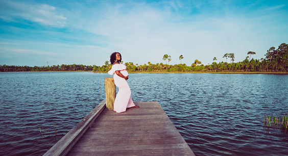 Beautiful pregnant African American woman stands on a scenic dock over the water