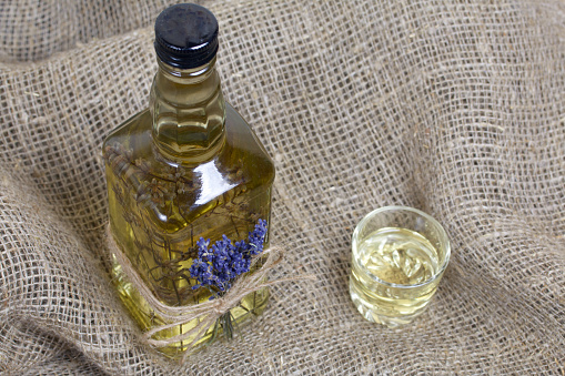 A bottle of lavender tincture. A bouquet of lavender is tied to the bottle. Nearby is a glass of drink. On coarse linen. Close-up shot.