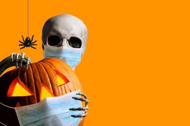 A skeleton wraps its hands around and peeks out from behind an illuminated jack o'lantern as a spider hangs from its web isolated against an orange background.  Both the skeleton and the jack o'lantern wear a protective face mask in the time of Covid-19.