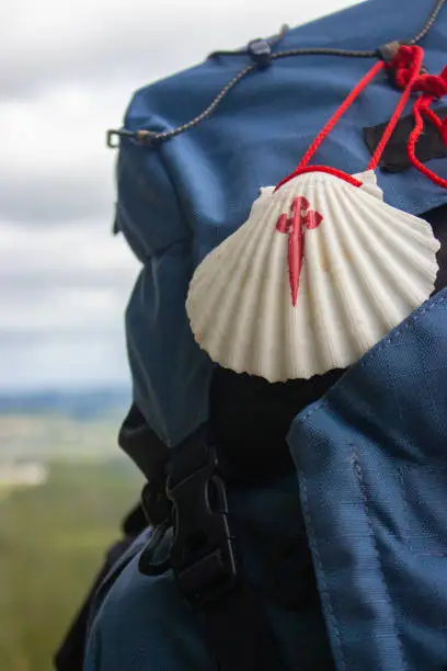 Photo of Big backpack with pilgrim shell on mountain background. Pilgrimage concept. Pilgrim on Camino de Santiago road. Tourist with rucksack on beautiful landscape background. Religious travel.