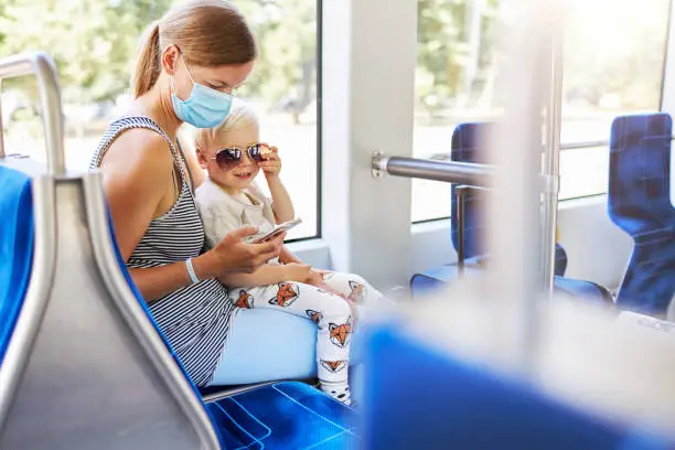 A young mother is sitting with her little son on her lap on the train while she is wearing a face mask and the toddler is playing with sunglasses, photographed in high resolution with copy space