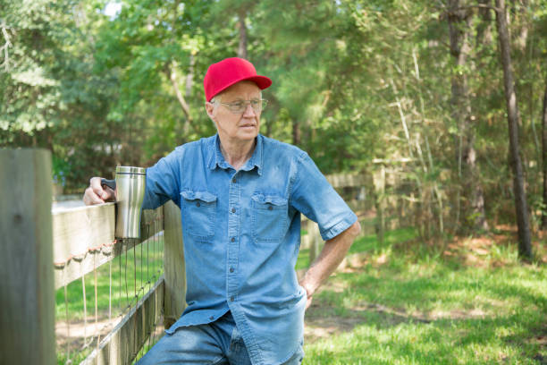 senior mann mit blick über seinen zaun.  er hält einen wiederverwendbaren kaffeebecher in der welt. - baseball cap old red caucasian stock-fotos und bilder