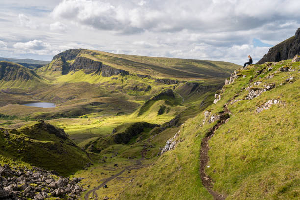 Side view of teeange girl sitting on rock, looking at the view of  Prison rocky outcrop in the Quiraing, Isle of Skye, Scotland. Beautiful area of grassy mountains, hidden lochs and steep cliffs stock photo