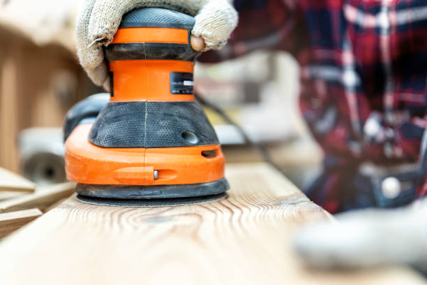 Closeup professional carpenter hand grinding raw wood plank with orbital sander electric machine in carpentry diy workshop. Detail of furniture restoration renewal. Power tool and equipment concept stock photo