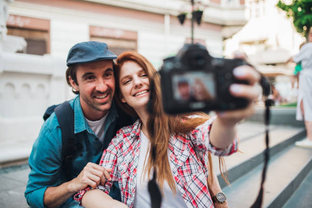 istock Happy couple on vacation sitting on the street making selfies 1272010607