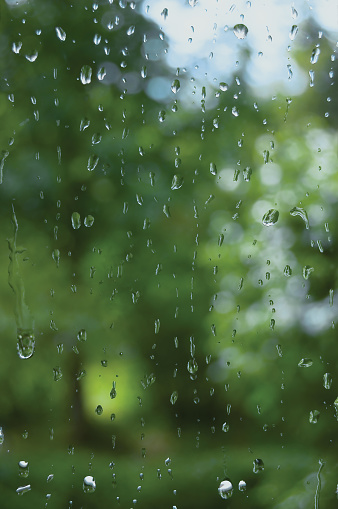 Rainy summer day, raindrops on window glass, rain droplets macro closeup, large detailed vertical copy space background, green trees, blue sky, gentle bokeh