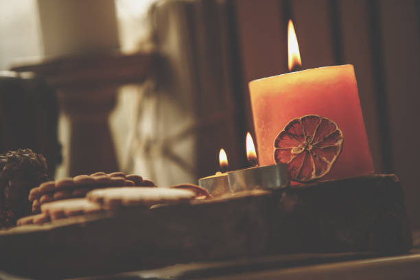 Low angle view of three lit candles in close up composition set on wooden board with tea cookies on the side and blurred background.