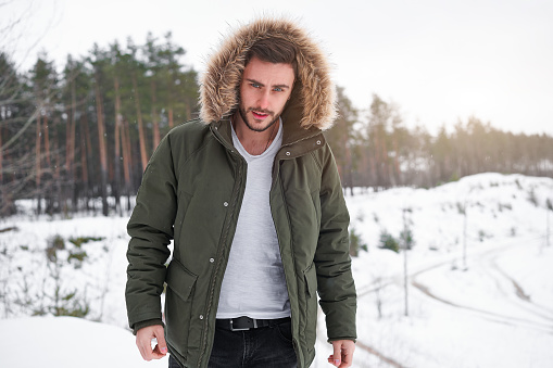 Handsome bearded young caucasian man standing outdoors fur hood in winter season forest. Attractive stylish european guy walking snowy christmas woodland Season holiday leisure