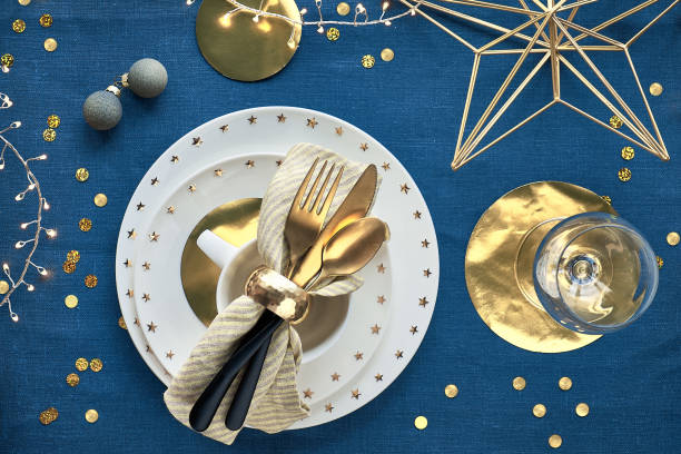 istock Christmas table setup with white plate and golden utensils and gilded decorations. Flat lay, top view on dark blue linen textile background. Xmas lights garland. 1272017460