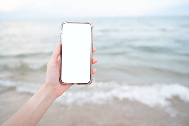istock Woman hand holding mobile phone with blank white screen mockup on the beach. 1272036285