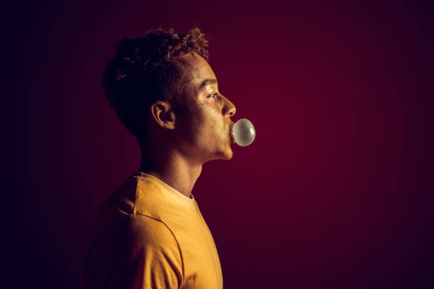 Young man standing isolated on dark red background in yellow lighting blowing bubble with gum pensive side view close-up copy space for text or product