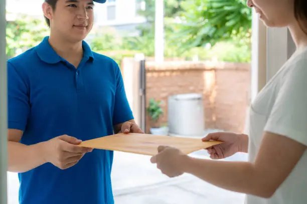 Asian delivery young man in blue uniform smile and holding letter or document envelope in front house and Asian woman accepting a delivery of envelope from deliveryman. Advertising, Business, Transportation Concept.