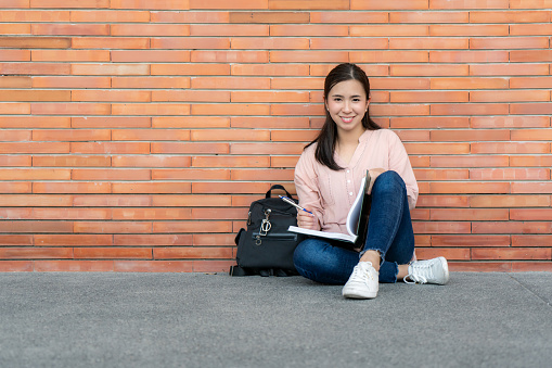Asian smiling woman student holding book posing on brick background in campus. Happy teen girl high school student outdoors. Education, Learning, Student, Campus, University, Lifestyle concept.