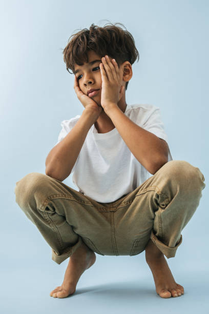 Thoughtful tired indian asian kid sitting low, knees up, hands on cheeks Thoughtful tired barefoot indian asian kid sitting low, knees up, hands on cheeks. Over blue background. indian boy barefoot stock pictures, royalty-free photos & images