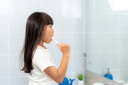 Cute Asian primary school girl brushing teeth and looking in mirror in bathroom at home. The morning school routine for day in the life getting ready for school.